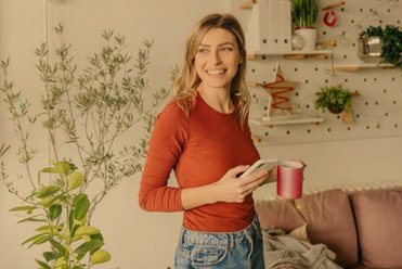 Junge Frau in ihrem Wohnzimmer mit Handy und Kaffeetasse in der Hand