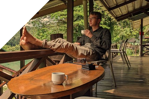 Ein junger Mann sitzt entspannt auf einer Terrasse und hält eine Tasse Kaffee in der Hand.