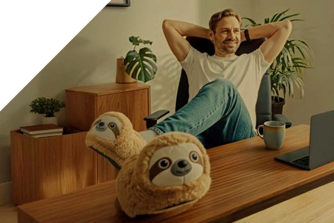 Mann lehnt sich entspannt im Schreibtischstuhl zurück  und streckt seine Füsse mit Faultierpantoffeln auf den Schreibtisch